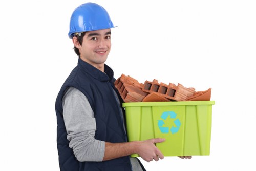 A young man wearing a blue hard hat and navy blue work vest, with a light gray long-sleeved shirt underneath, is holding a lime green recycling bin marked with a blue recycling symbol. Inside the bin are several red clay bricks stacked neatly, with rough textured surfaces and characteristic rectangular holes. The bricks' warm terracotta color contrasts with the smooth, vibrant finish of the bin. The man is standing against a plain white background, smiling subtly and positioned slightly to the right of the image. The scene appears to be studio-lit, with even lighting highlighting the textures of the bricks and the reflective surface of the bin. This context suggests a professional waste removal or recycling service, consistent with offerings by companies like Office Clearance Regents Park, serving areas within or near central London, including the Regents Park postcode district.