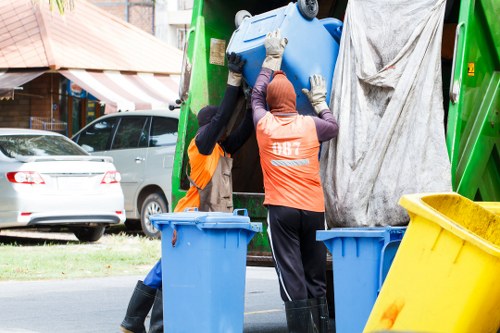 Two waste management workers are seen loading a large blue plastic bin into a green rubbish collection vehicle on a residential street. One worker, dressed in an orange high-visibility vest and black trousers, is on the left while the other, wearing an orange vest with branding on the back and a head covering, stands on the right. The vehicle is parked close to the pavement, and the workers are lifting the bin over its side. In the background, there are parked cars, including a silver vehicle, and a row of houses with brick facades and striped awnings. A yellow waste container is positioned on the pavement, indicating ongoing rubbish collection or clearance. The scene takes place during daylight hours in a suburban area, with a focus on professional waste removal activities typical for companies like officeclearanceregentspark.co.uk serving the Regents Park region.