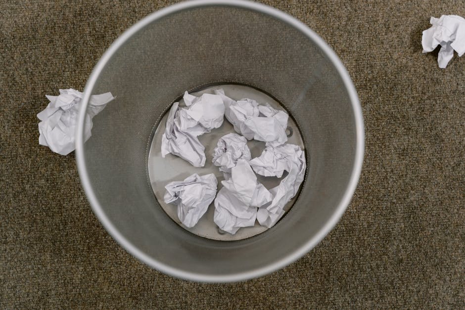 Two large, clear plastic bags filled with discarded waste are positioned on a paved sidewalk in front of a dark wooden fence and dense foliage. The bags appear to contain mixed rubbish, possibly inclu