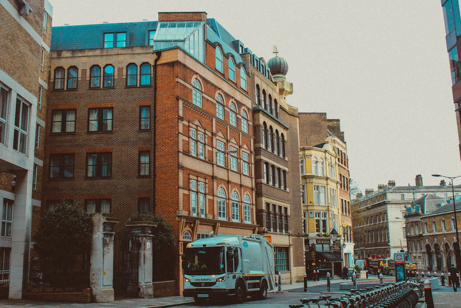 A small waste collection truck parked on a city street in front of an orange-brick residential building with multiple windows and black metal balconies. The truck has a white body with some visible re