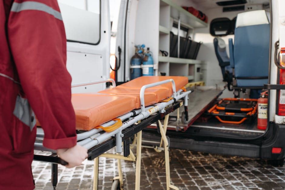 The image shows the interior of a white emergency response or medical transport vehicle, with a focus on a wheeled stretcher positioned near the open rear door. The stretcher has an orange, padded mat