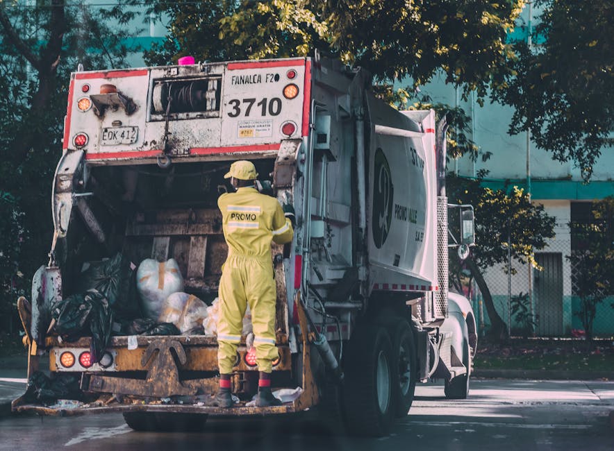 A waste collection truck is parked on a street with its rear compartment open, revealing large black garbage bags filled with waste and recycling material. The truck is predominantly white with a red