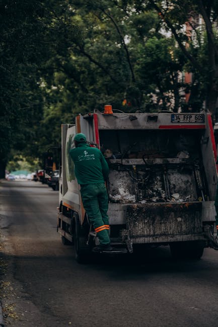 A waste collection truck is parked on a street with its rear compartment open, revealing large black garbage bags filled with waste and recycling material. The truck is predominantly white with a red