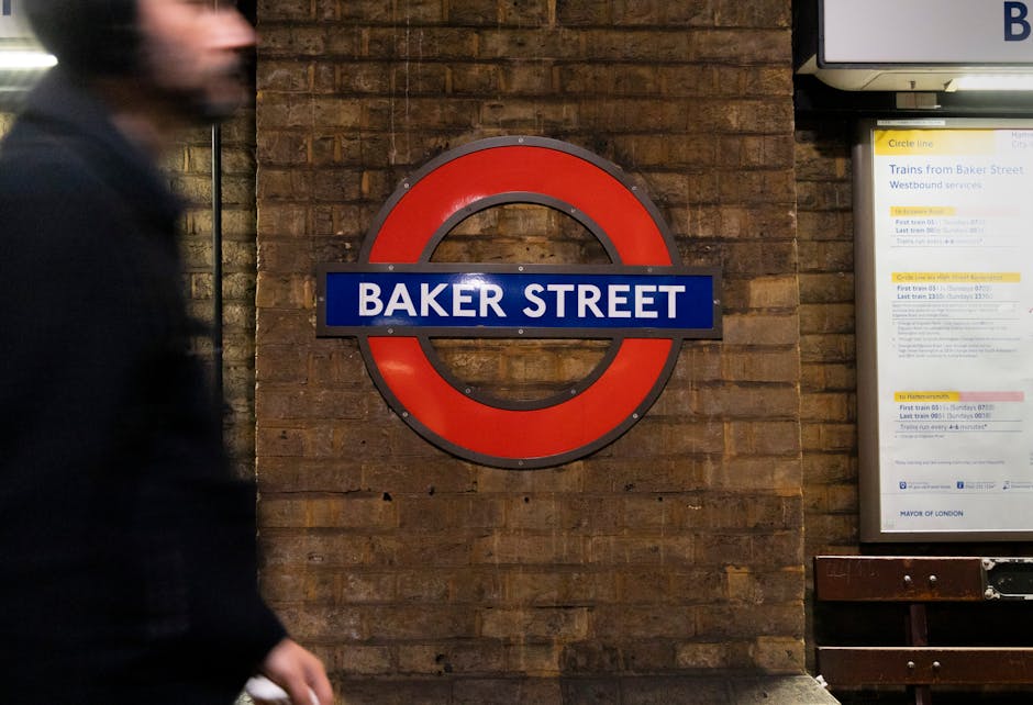 A section of an indoor urban setting featuring a circular London Underground station sign mounted on a brown brick wall. The sign displays the text 'BAKER STREET' in white uppercase letters on a blue 