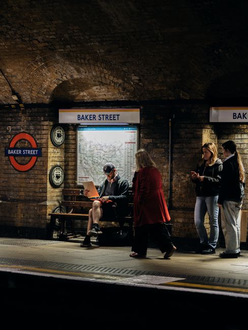 A section of an indoor urban setting featuring a circular London Underground station sign mounted on a brown brick wall. The sign displays the text 'BAKER STREET' in white uppercase letters on a blue 