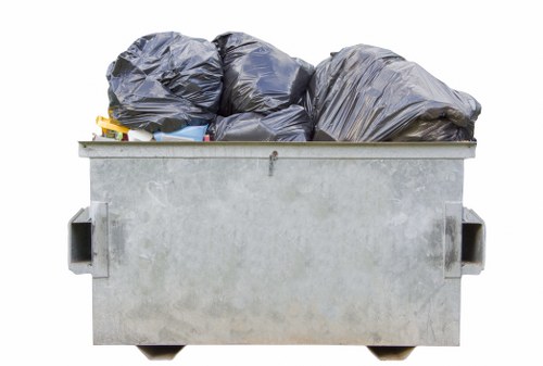 A large metal skip filled with multiple black rubbish bags and loose debris, situated outdoors on a paved surface, with a plain background. The bags appear to contain household or office waste, and they are tightly packed, with some bags leaning slightly to one side. The metal skip has a utilitarian, slightly weathered appearance, with visible handles on each side. Surrounding environment is minimal, but the skip's setting suggests an urban or suburban location, possibly in Regents Park or nearby area. The scene aligns with rubbish removal services offered by Office Clearance Regents Park, illustrating the typical scope of waste collection and disposal tasks in the local area. Natural daylight illuminates the scene, emphasizing the textures of the bags and the metal surface of the skip, which is ready for waste collection in support of efficient office or property clearance activities.