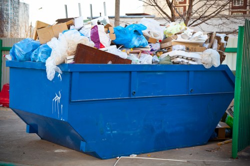 A large blue metal skip filled with mixed waste materials is positioned on a paved surface outdoors, adjacent to a green metal fence. The skip contains various types of rubbish, including cardboard boxes, white plastic packaging, blue plastic bags, and other debris, some of which are partially hanging over the sides. In the background, there are blurred trees and distant buildings, indicating an urban or residential setting. The overall scene suggests a waste collection point in the vicinity of a property requiring rubbish removal, consistent with services provided by Office Clearance Regents Park. The image highlights the need for professional rubbish disposal and clearance solutions in the borough, emphasizing practical waste management in the local area.
