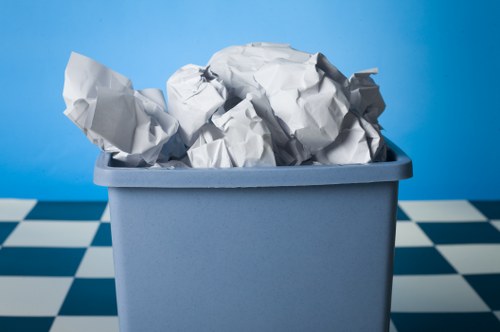 A close-up of a light blue plastic rubbish bin filled with crumpled white paper waste, with some paper sheets protruding over the top edge. The bin is situated outdoors on a tiled pavement with a black and white checkered pattern, set against a plain blue backdrop. The crumpled paper appears to be used office or household waste, with visible folds and creases on the sheets. The environment suggests a typical urban setting within Regent's Park or nearby London areas, consistent with local rubbish removal services offered by Office Clearance Regents Park. The scene captures a straightforward view of waste ready for collection, emphasizing the importance of proper rubbish disposal and clearance services in the local community.