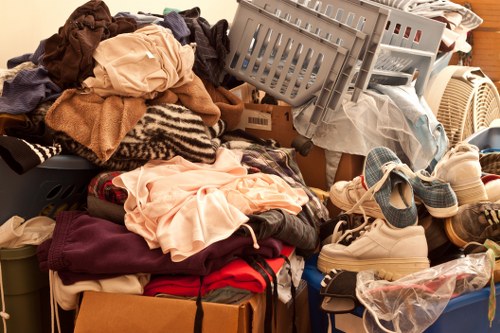 An indoor scene showing a large assortment of cluttered household textiles and miscellaneous items scattered across a surface, including piles of clothes in various colours and fabrics, some crumpled and folded, with textiles ranging from dark maroon and beige to light pink and patterned fabrics. There are also several plastic storage baskets, some tilted and overflowing with fabric or paper, and a cardboard box partially visible underneath the pile. A pair of white sneakers with blue laces and a plastic bag containing more items are visible among the clutter. The background includes a portion of a wooden surface and a heating radiator. This scene exemplifies typical household waste that a rubbish removal service such as Office Clearance Regents Park might manage as part of an office clearance or domestic decluttering project in the local London area, particularly near the Regent’s Park postcode zone, with the clutter spread across a flat surface in a room or storage area.